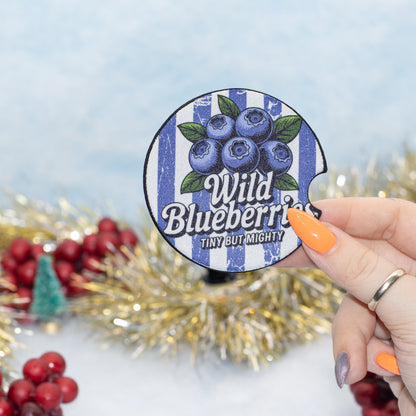 A hand holds a round blue-and-white striped coaster featuring blueberries and the text 'Wild Blueberries' and 'TINY BUT MIGHTY'. The background is a festive holiday setting with gold tinsel, red berries, and a soft light blue backdrop.