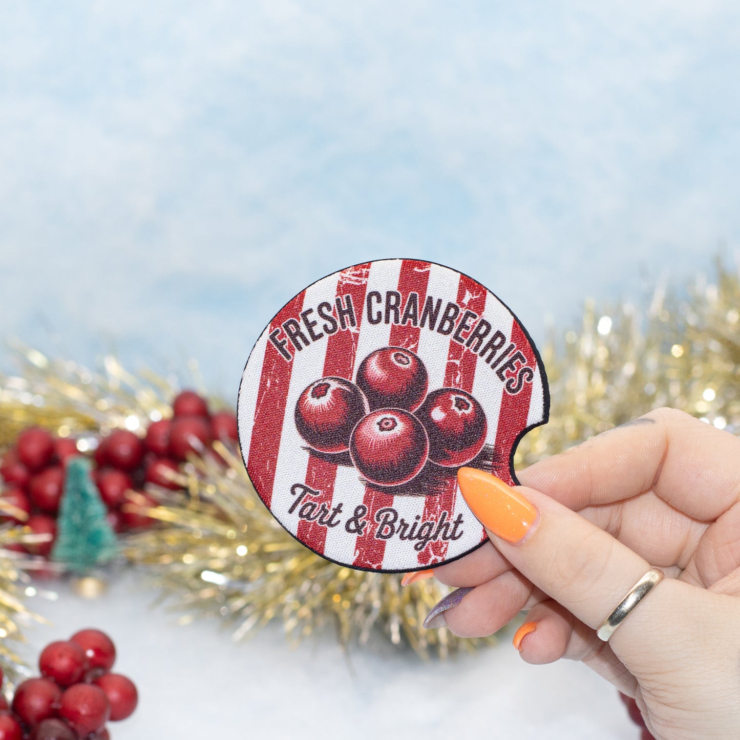A hand holds a round, car coaster featuring red and white vertical stripes, black text reading 'FRESH CRANBERRIES Tart & Bright,' and illustrations of three cranberries. The background is a soft light blue with blurred holiday decorations, including red berries, gold tinsel, and a small green tree.