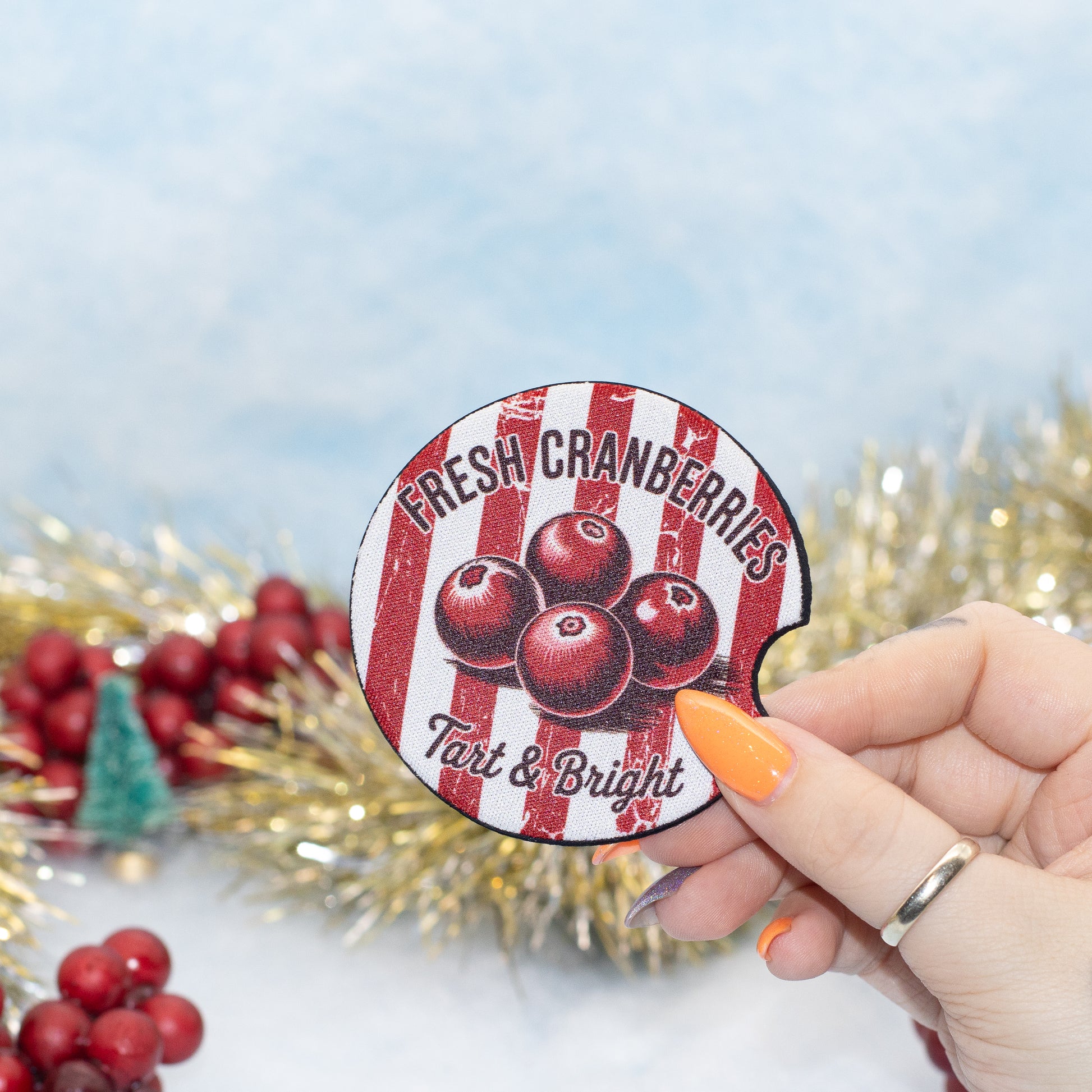 A hand holds a round, car coaster featuring red and white vertical stripes, black text reading 'FRESH CRANBERRIES Tart & Bright,' and illustrations of three cranberries. The background is a soft light blue with blurred holiday decorations, including red berries, gold tinsel, and a small green tree.