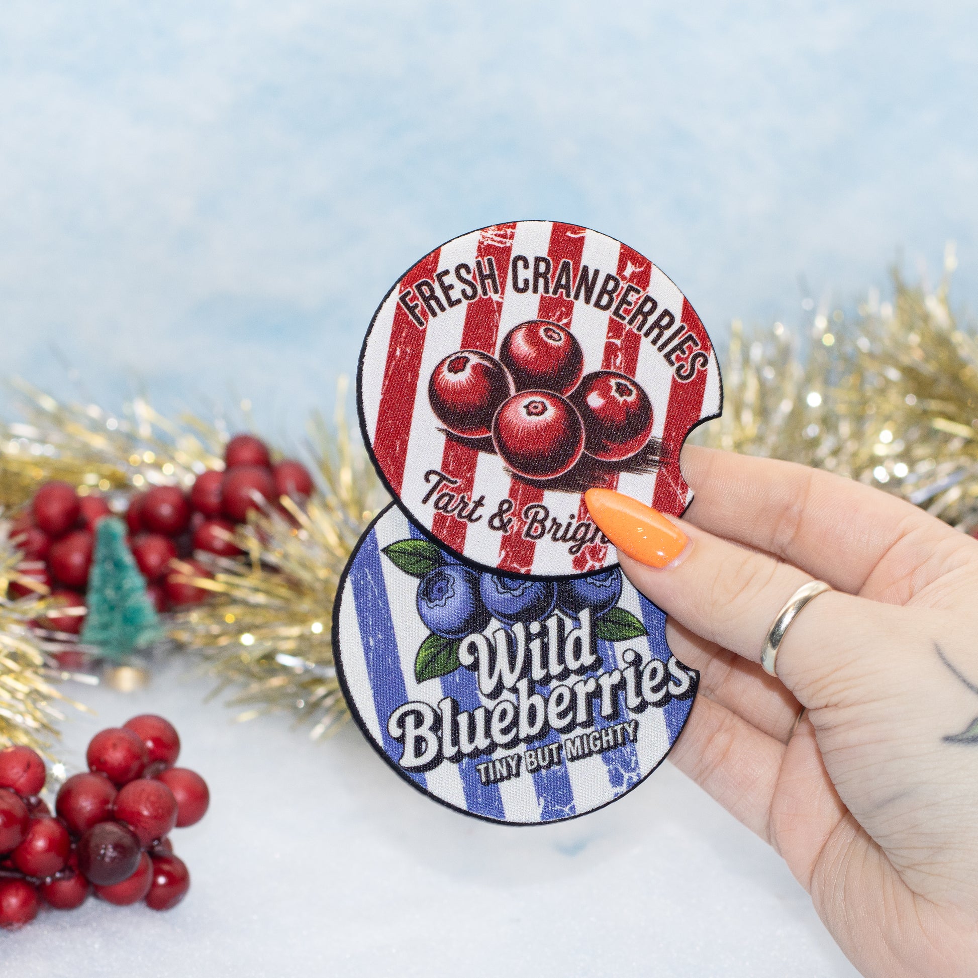 A hand holds two round, car coasters featuring berry-themed designs. The top sticker reads 'Fresh Cranberries' with red cranberries on red-and-white stripes, and the bottom sticker reads 'Wild Blueberries' with blueberries on blue-and-white stripes.