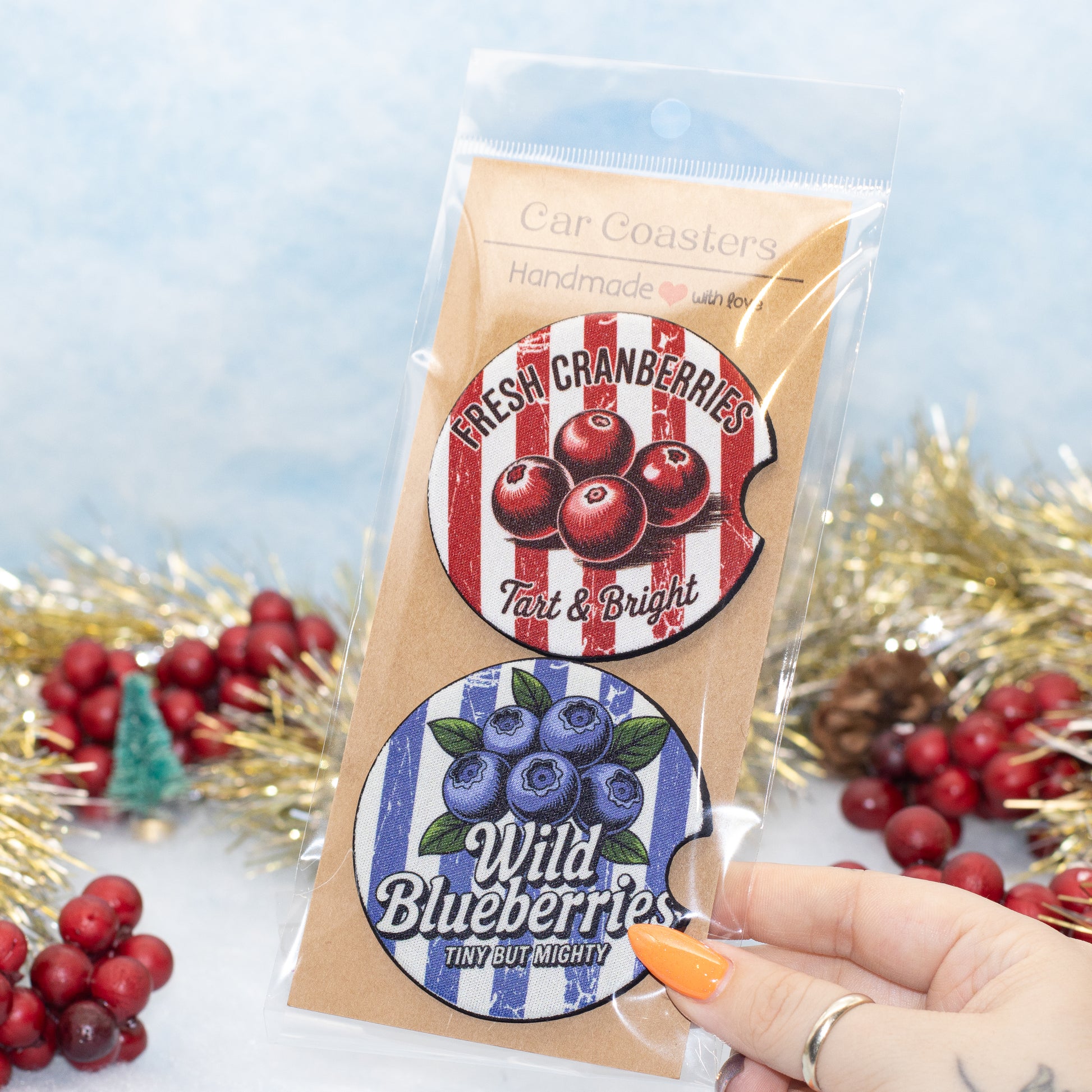 A hand holds a clear plastic bag containing two round car coasters. The top coaster is labeled 'Fresh Cranberries' with red berries on a red-and-white striped background, and the bottom coaster is labeled 'Wild Blueberries' with blue berries on a blue-and-white striped background.