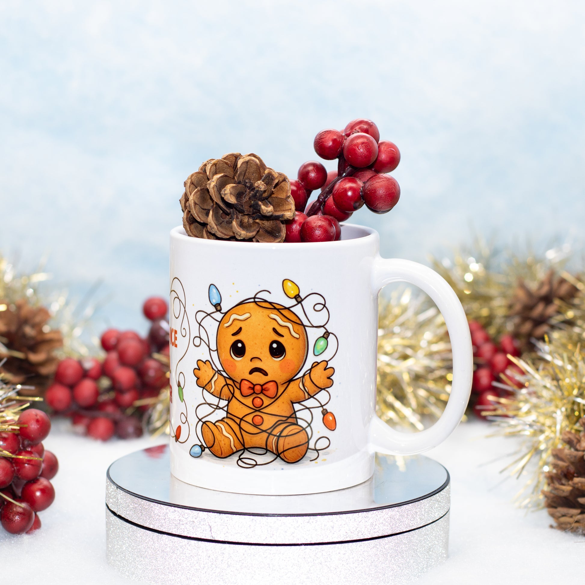 A white ceramic mug with a cute gingerbread character illustration, styled with a pinecone and red berries on top, set against a festive holiday background with gold tinsel.