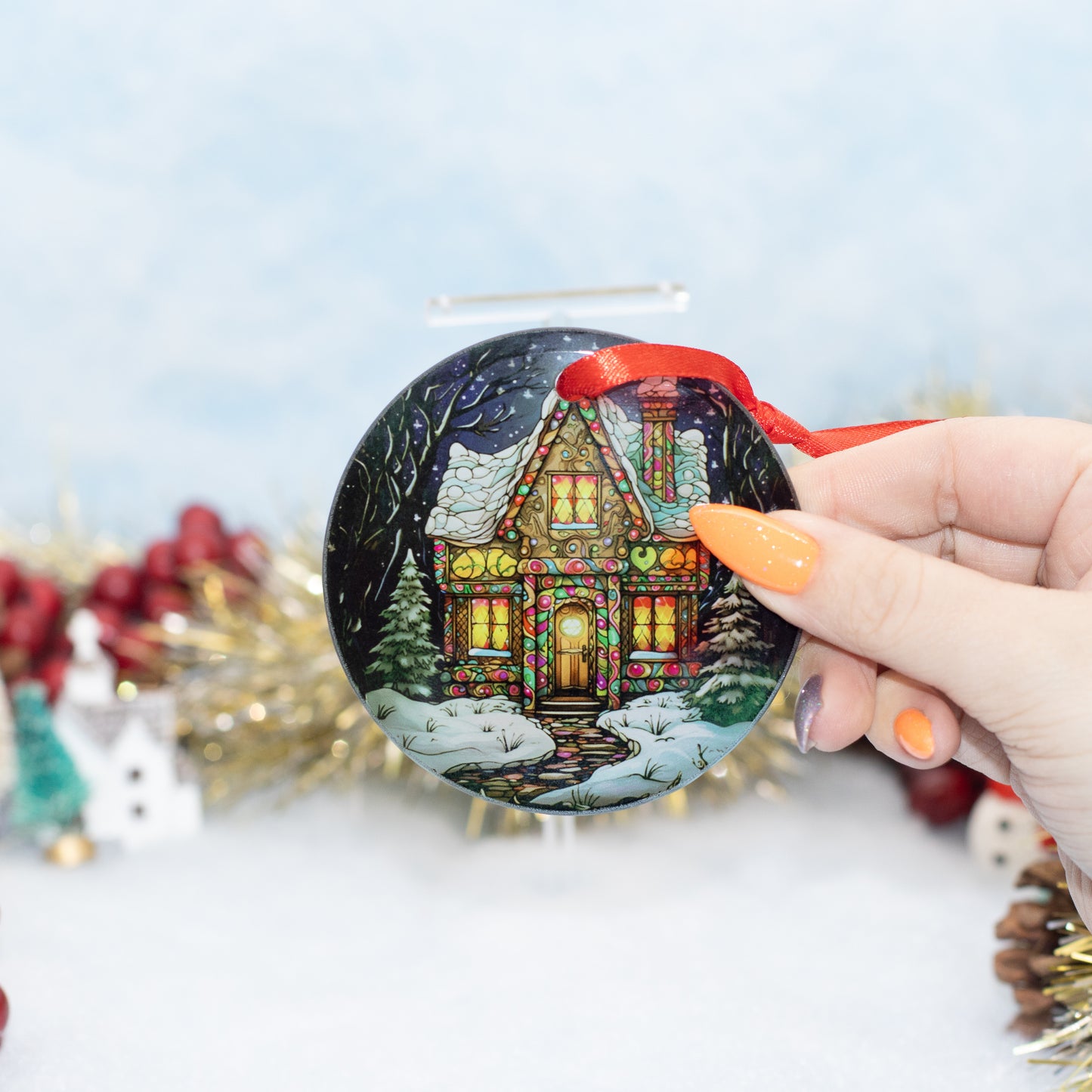 A hand holds a round Christmas ornament featuring a festive stained-glass-style house with snow, green trees, and a red hanging ribbon. The background is softly blurred with holiday decorations.