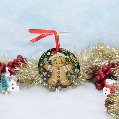 A round Christmas ornament featuring a gingerbread man on a dark patterned background, hanging from a red ribbon. The ornament is styled in a festive holiday setting with gold tinsel, red berries, pinecones, and a small snowman figure against a soft light blue background.