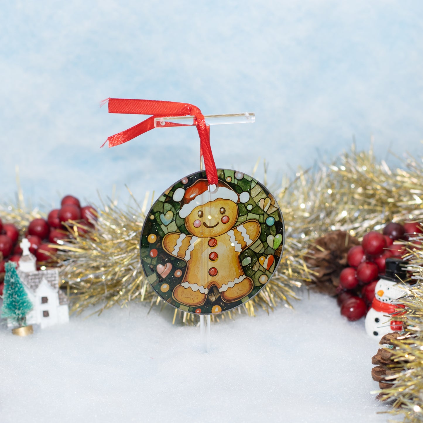 A round Christmas ornament featuring a  gingerbread man on a decorated background, hanging from a red ribbon on a clear stand. The ornament is styled with gold tinsel, red berries, pinecones, and a small snowman figure against a soft blue and white background.