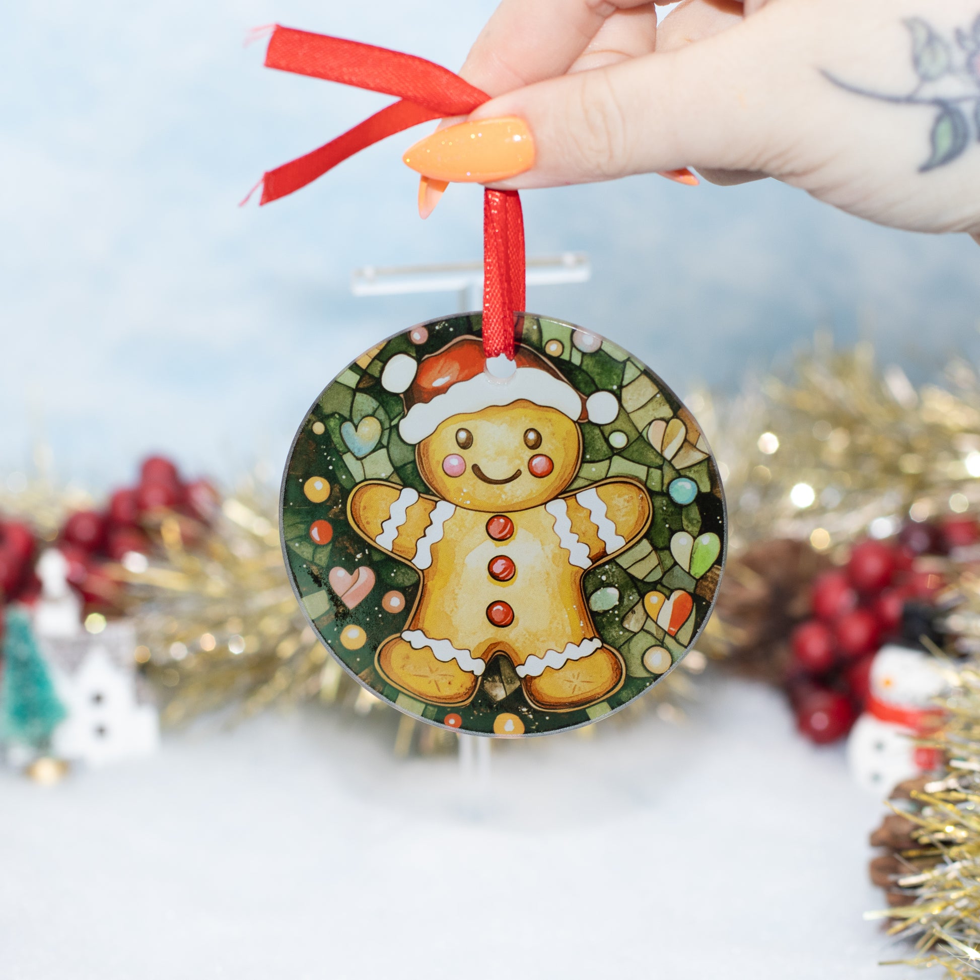 A round Christmas ornament featuring a painted gingerbread person on a dark green patterned background, hanging from a red ribbon held by a hand with orange nail polish.