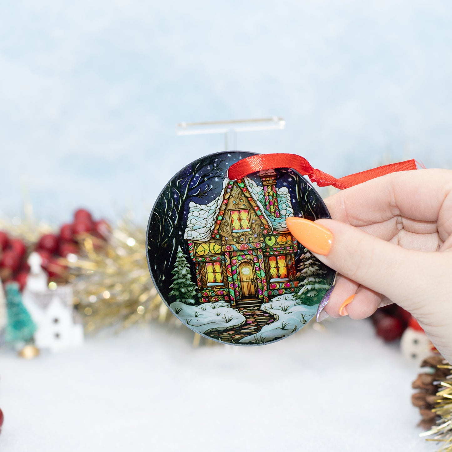 A hand holds a round Christmas ornament featuring a painted winter cabin scene with snow, trees, and warm glowing windows. The ornament has a red ribbon loop and is photographed against a softly blurred festive background with tinsel and holiday decor.