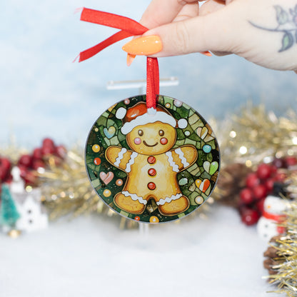 A round Christmas ornament featuring a gingerbread man illustration, held by a hand with orange nail polish. The ornament has a red ribbon loop and is shown against a festive, blurred holiday background with gold tinsel and red berries.