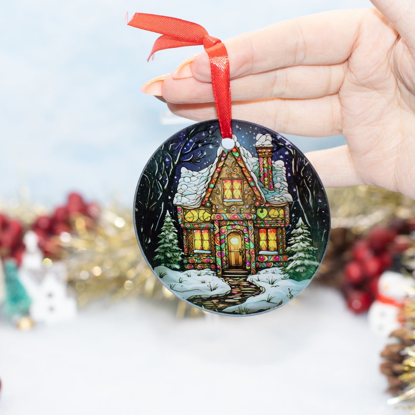 A hand holds a round Christmas ornament featuring a painted cozy house at night with snow, evergreen trees, and warm glowing windows. The ornament has a red ribbon loop and is photographed against a softly blurred festive background with gold tinsel and red berries.