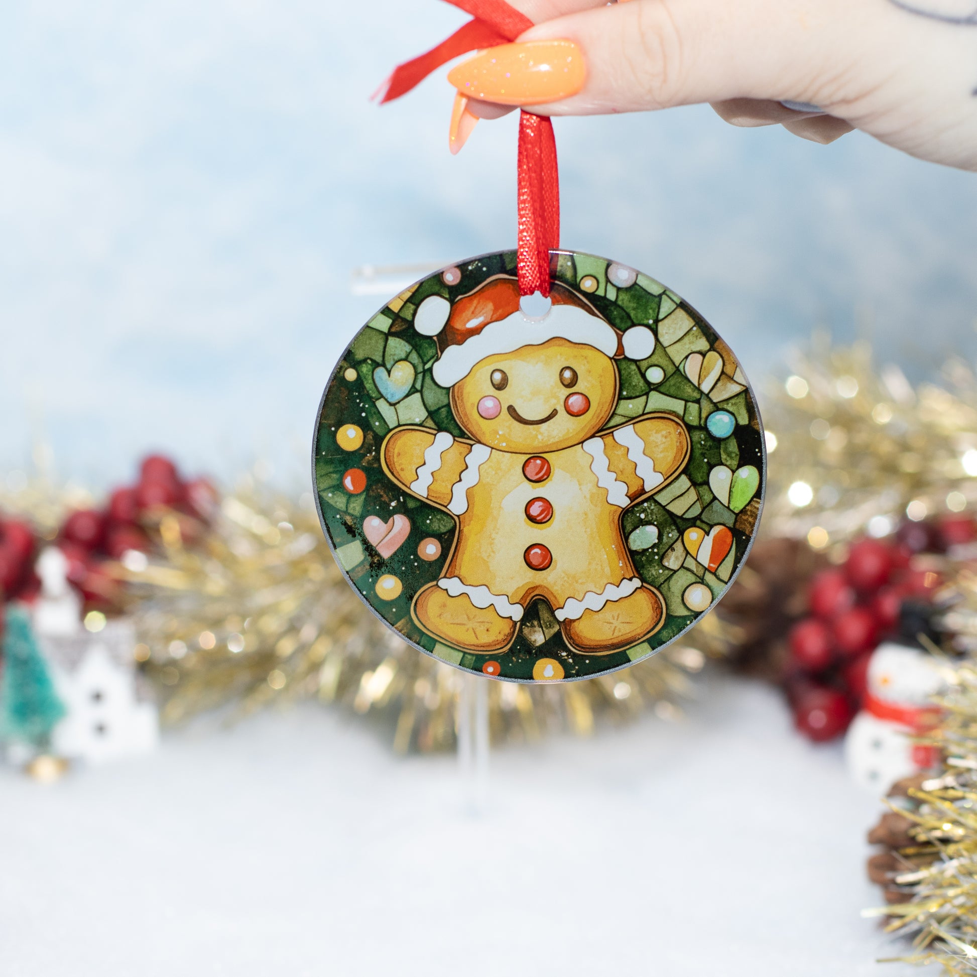 A round Christmas ornament featuring a gingerbread man illustration, held by a hand with orange nail polish. The ornament has a red hanging ribbon and is set against a festive blurred background with gold tinsel and red berries.