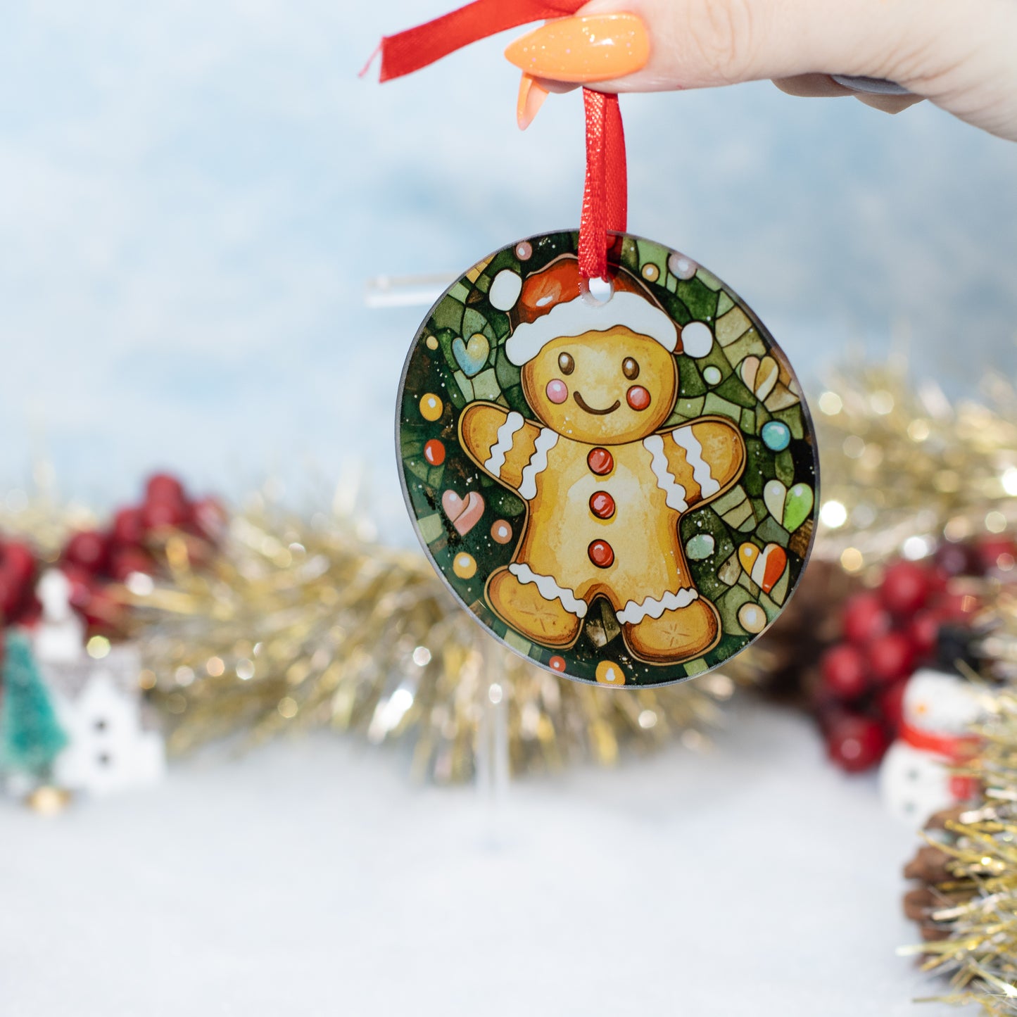 A hand holds a round Christmas ornament featuring a painted gingerbread man on a decorated tree background. The ornament has a red ribbon loop and is photographed against a softly blurred holiday backdrop with tinsel and red berries.