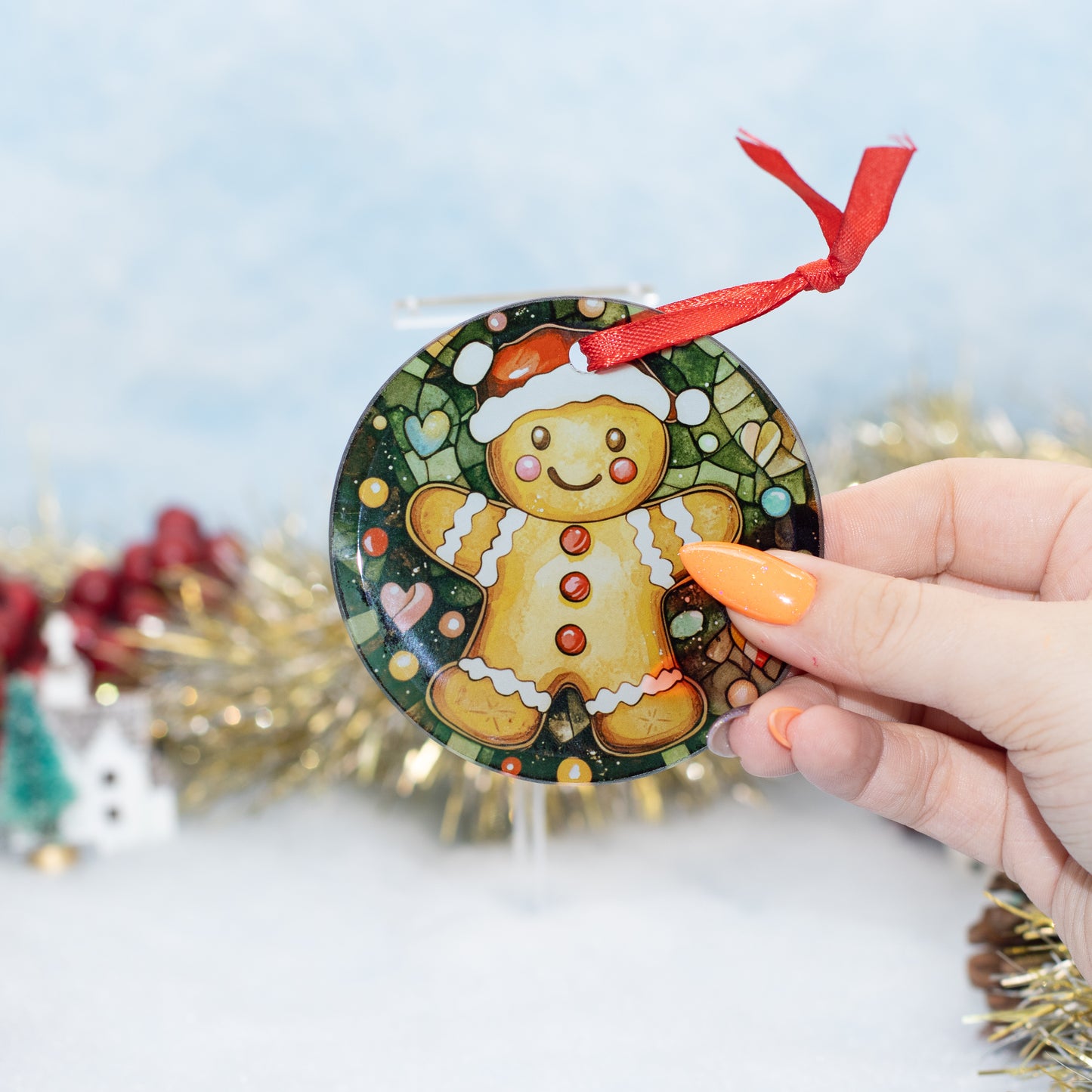 A hand holds a round Christmas ornament featuring a gingerbread man in a stained-glass style. The ornament has a red ribbon loop at the top and is shown against a festive, blurred background with gold tinsel and a light blue backdrop.