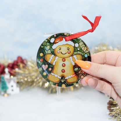 A hand holds a round Christmas ornament featuring a gingerbread man in a stained-glass style. The ornament has a red ribbon loop at the top and is shown against a festive, blurred background with gold tinsel and a light blue backdrop.