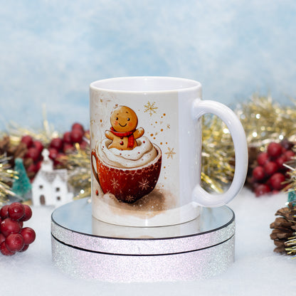 A white ceramic mug with a festive Christmas illustration of a gingerbread man on whipped cream, displayed on a round silver glitter pedestal with holiday decorations in the background.