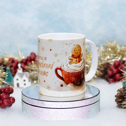 A white ceramic mug with a festive Christmas design, featuring a gingerbread man on a cup of hot chocolate with whipped cream and the text 'MUG FULL OF Gingerbread dreams'. The mug is displayed on a round silver glitter pedestal with holiday decorations in the background.