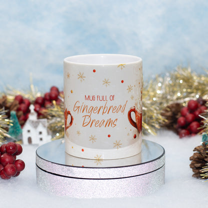 A white ceramic mug with festive gingerbread-themed illustrations and the text 'MUG FULL OF Gingerbread Dreams' in orange, displayed on a round mirrored stand. The background is decorated with gold tinsel, red berries, and a soft blue textured backdrop.