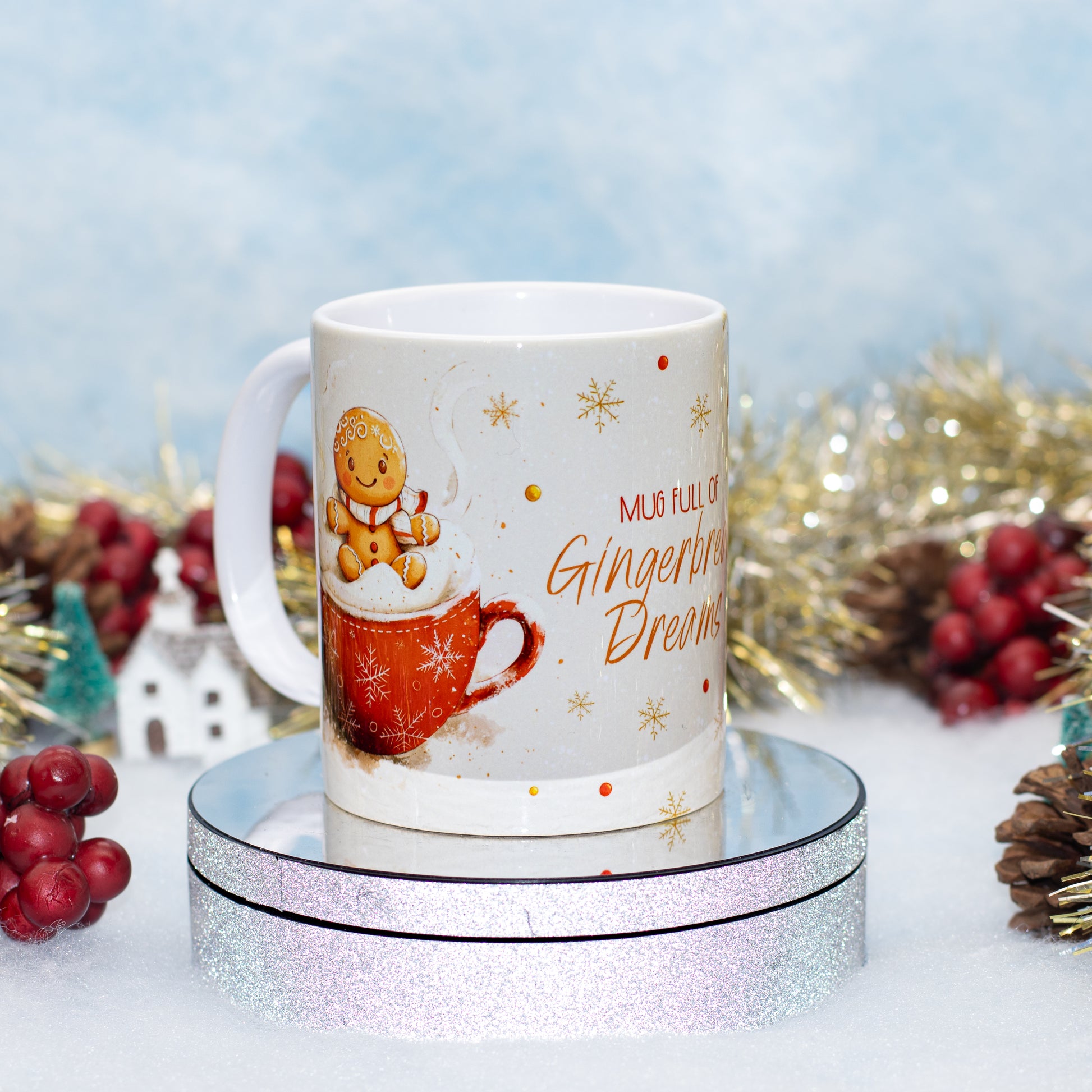 A white ceramic mug with a gingerbread-themed holiday design, featuring a gingerbread man sitting on a red mug with whipped cream and the text 'MUG FULL OF Gingerbread Dreams'. The mug is displayed on a round silver glitter pedestal with festive tinsel and red berries in the background.