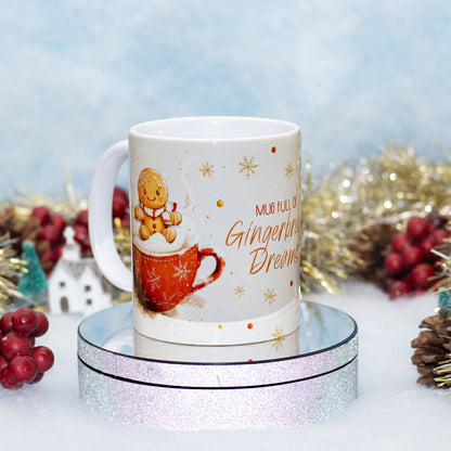 A white ceramic mug with a gingerbread-themed holiday design, featuring a gingerbread man sitting on a red mug with whipped cream and the text 'MUG FULL OF Gingerbread Dreams'. The mug is displayed on a round silver glitter pedestal with festive tinsel and red berries in the background.