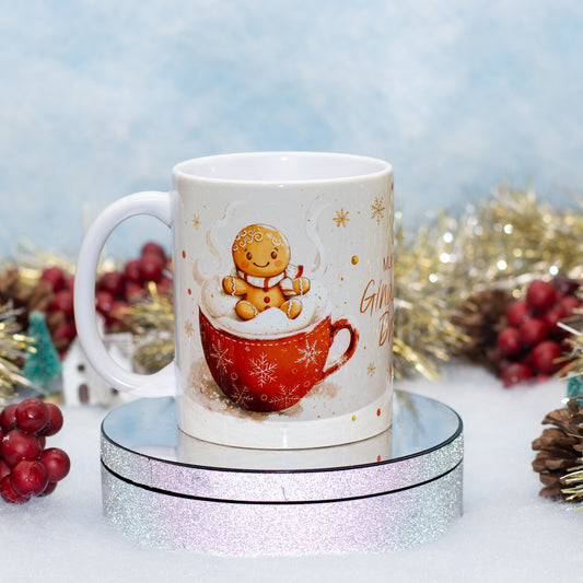 A white ceramic mug with a Christmas design, featuring a gingerbread man sitting in a red mug filled with whipped cream and hot cocoa. The mug is displayed on a round, glittery silver pedestal with festive holiday decorations in the background.