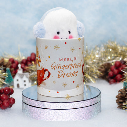 A white ceramic mug with a plush white character peeking out, decorated with gold snowflakes and the text 'MUG FULL OF Gingerbread Dreams.' The mug sits on a round mirrored display base against a snowy, festive background with red berries, gold tinsel, and pinecones.