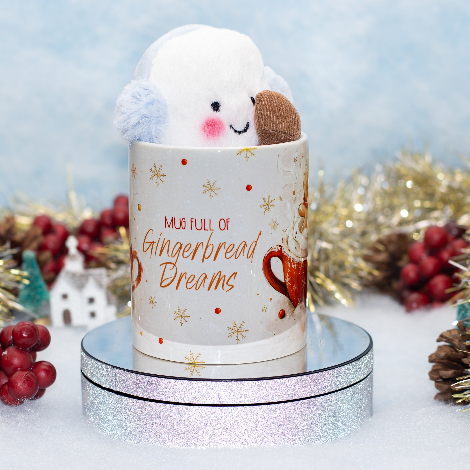 A white ceramic mug with gold snowflakes and red lettering reading 'MUG FULL OF Gingerbread Dreams,' topped with a white plush cloud character wearing a small blue ear warmer and holding a brown gingerbread cookie. The mug sits on a round silver glitter display base against a soft blue backdrop with holiday tinsel and red berries around it.