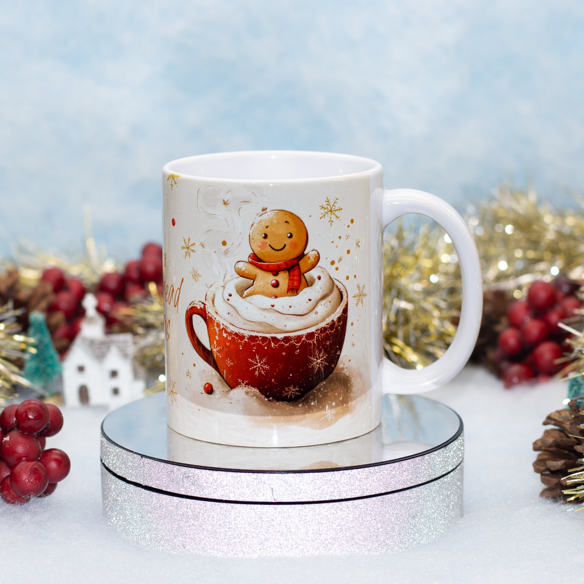 A white ceramic mug with a Christmas-themed illustration of a gingerbread man sitting in a red mug of hot cocoa with whipped cream, displayed on a round silver glitter stand with festive holiday decorations in the background.