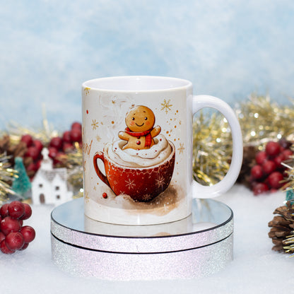 A white ceramic mug with a Christmas-themed illustration of a gingerbread man sitting in a red mug of hot cocoa with whipped cream, displayed on a round silver glitter stand with festive holiday decorations in the background.