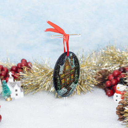 An oval Christmas ornament featuring a colorful illustration of a snowy house at night, hanging from a red ribbon on a clear stand. The ornament is styled on a white surface with gold tinsel, red berries, pinecones, and small holiday decorations in the background.
