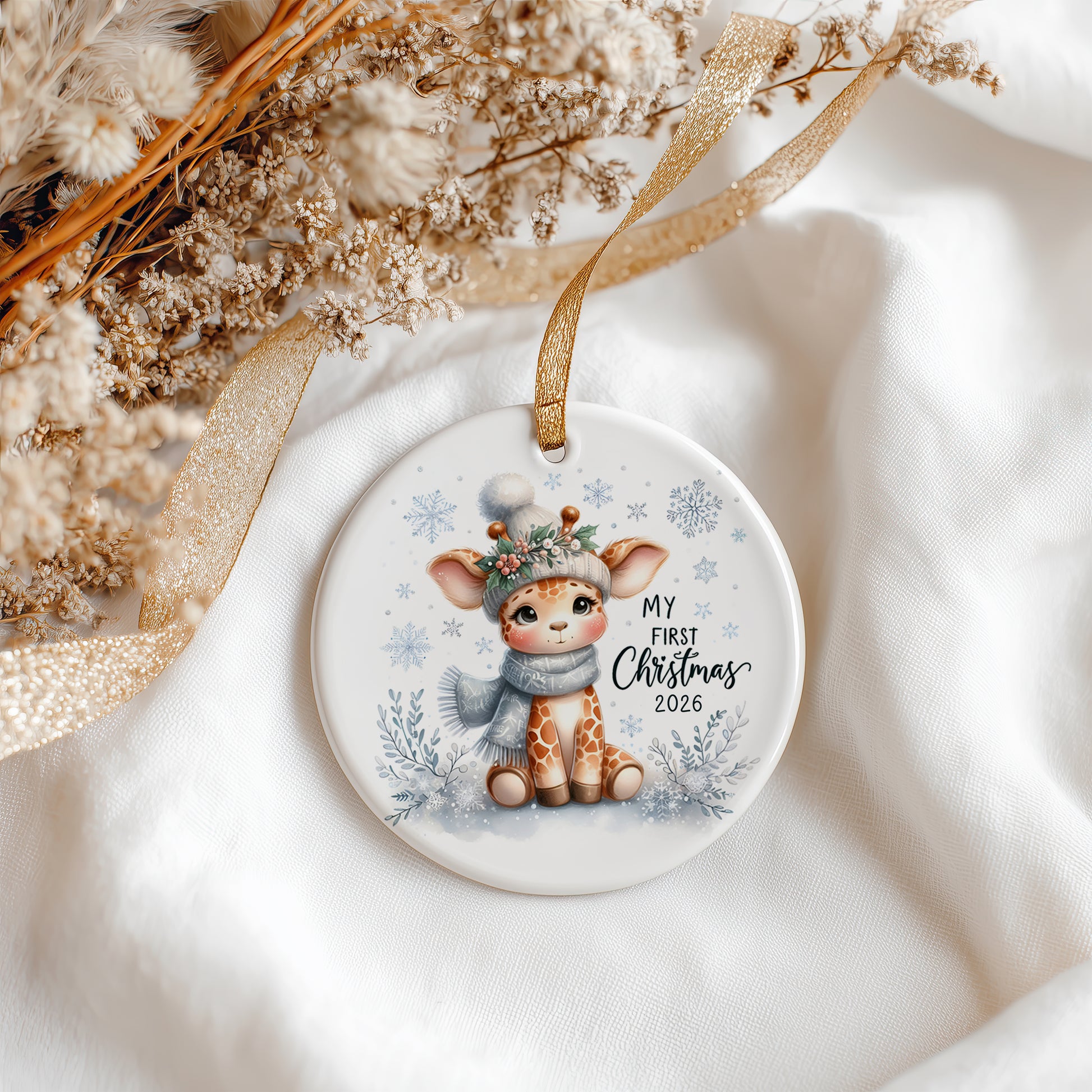 Round ceramic Christmas ornament featuring an illustrated baby deer wearing a green leafy hat and blue-gray scarf, with the text 'MY FIRST Christmas 2026' on a white background. The ornament hangs from a gold ribbon and is styled with dried flowers and a gold ribbon on white fabric.