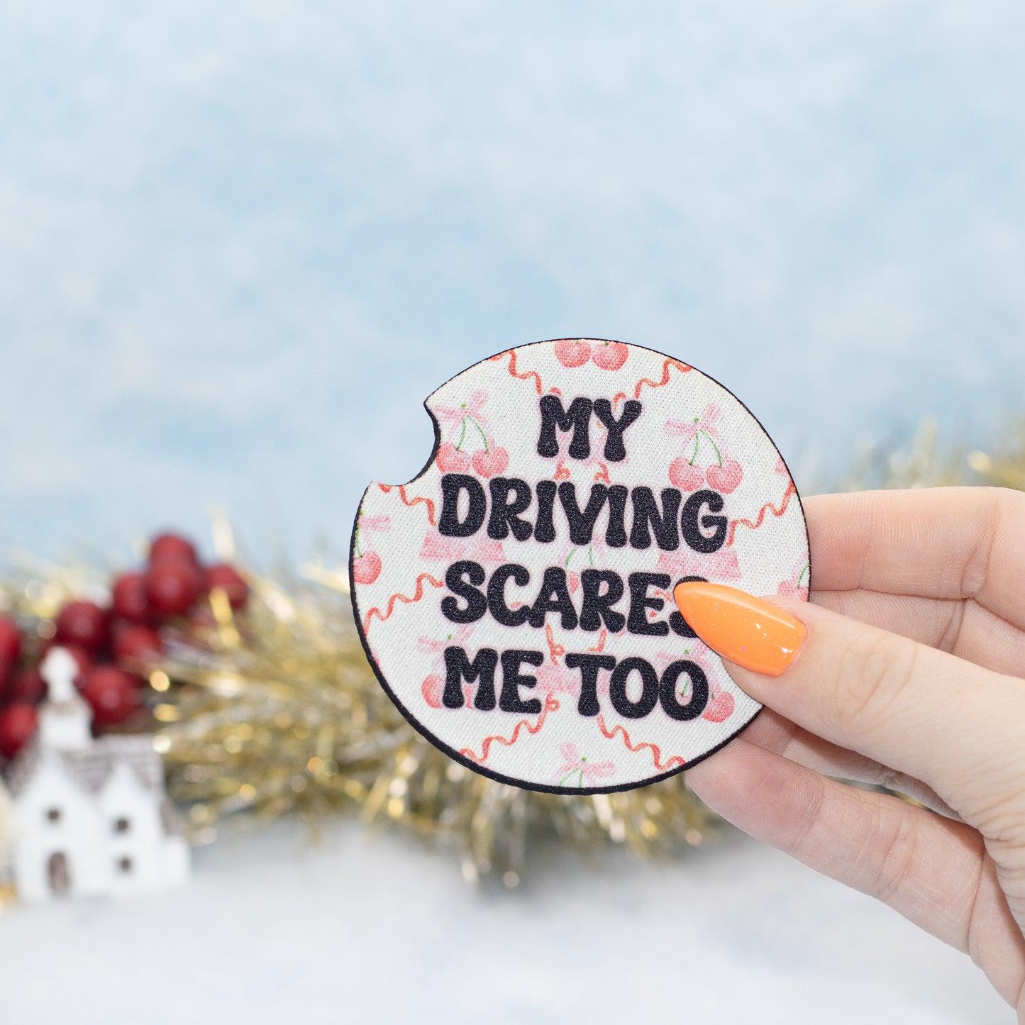 A hand hold a car coaster that reads 'MY DRIVING SCARE ME TOO' in black text on a white background with pink cherry illustrations. The background is a soft, out-of-focus light blue with gold tinsel and red berries.