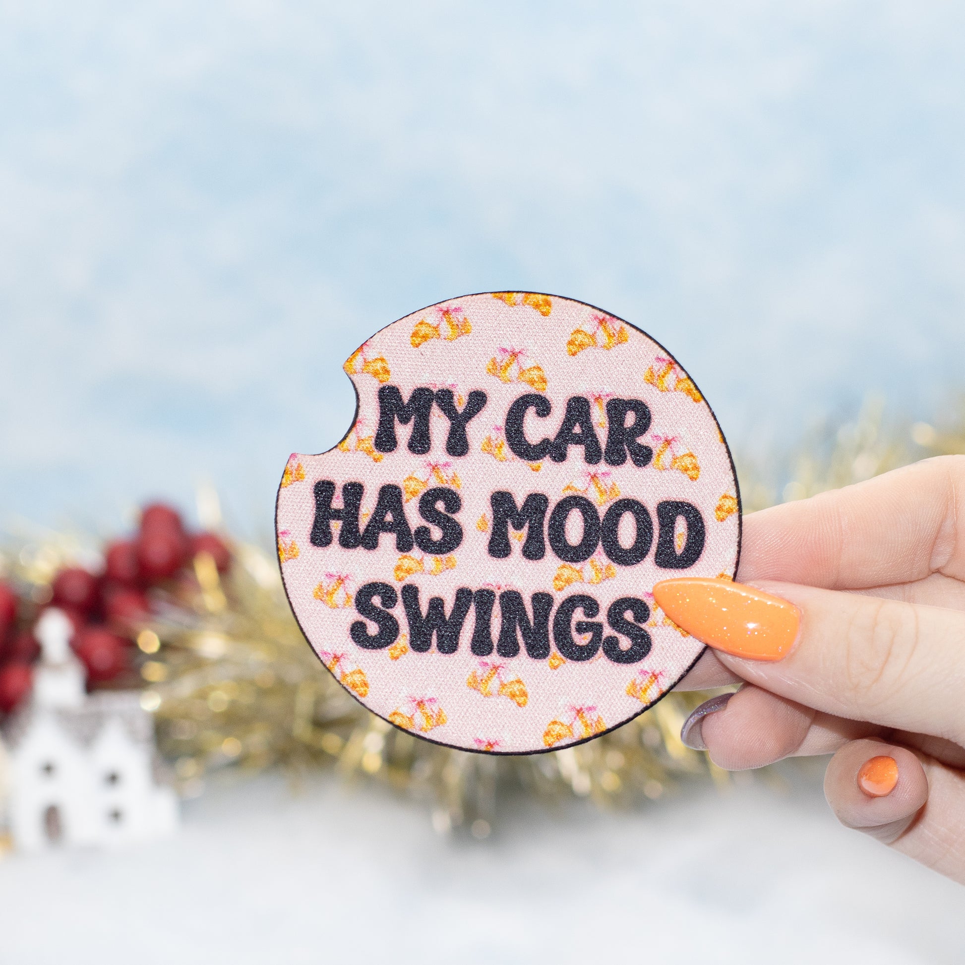 A hand holds a car coaster that reads 'MY CAR HAS MOOD SWINGS' in black text on a light pink background with small yellow and orange car icons. 