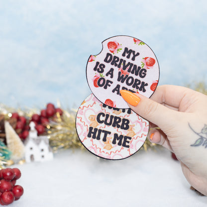 A hand hold two car coasters with black text reading "MY DRIVING IS A WORK OF ART" and "THAT CURB HIT ME" on a light pink strawberry-patterned background. The scene is styled with holiday decorations and a soft blue backdrop.
