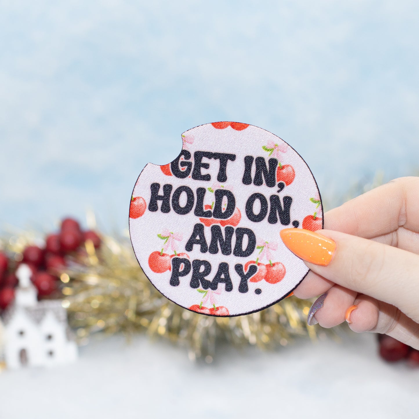 A hand holds a car coaster featuring the text 'GET IN, HOLD ON. AND PRAY.