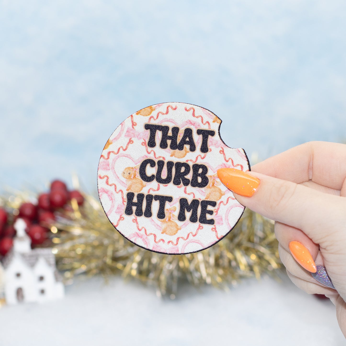 A hand hold a car coaster featuring the text 'THAT CURB HIT ME' in black letters over a pink and beige floral pattern. The background is a soft-focus holiday scene with gold tinsel and red berries.