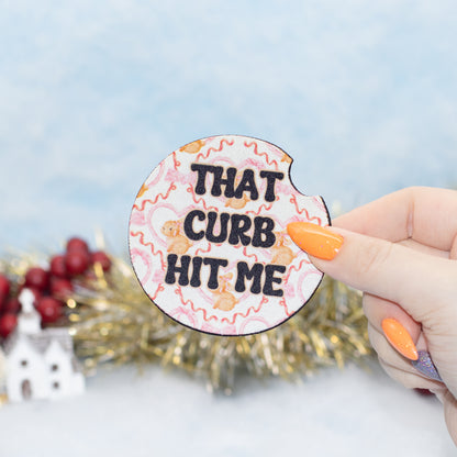 A hand hold a car coaster featuring the text 'THAT CURB HIT ME' in black letters over a pink and beige floral pattern. The background is a soft-focus holiday scene with gold tinsel and red berries.