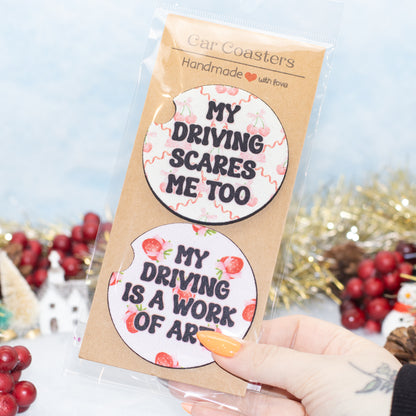 A hand holds a clear plastic bag containing two round car coasters with humorous driving-themed quotes on a kraft card backing. The background is decorated with red berries and gold tinsel.