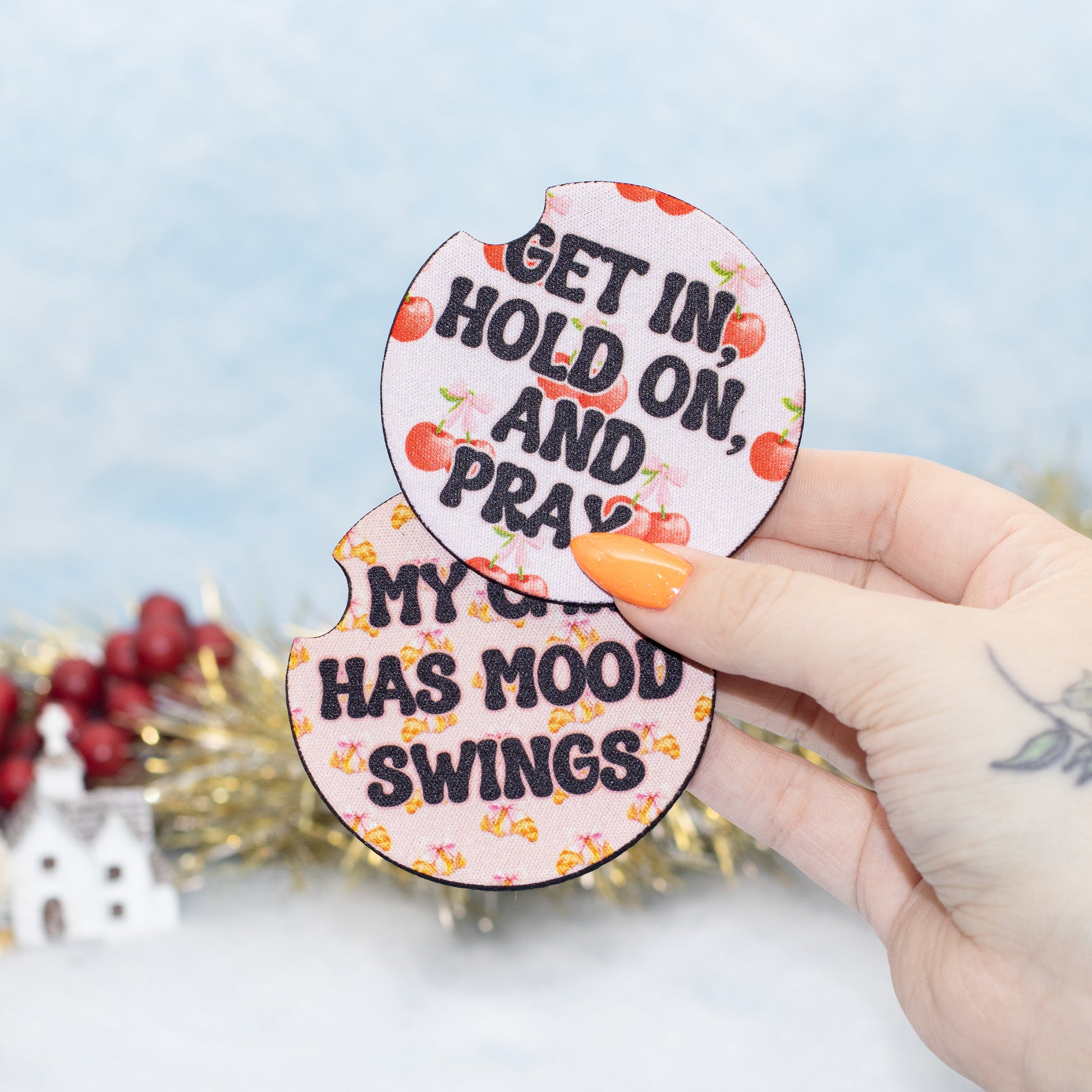 A hand holds two round, car coasters with cherry illustrations and text reading 'GET IN, HOLD ON, AND PRAY' and 'MY CAR HAS MOOD SWINGS'.