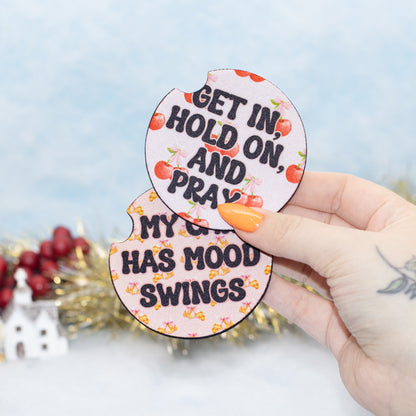 A hand holds two round, car coasters with cherry illustrations and text reading 'GET IN, HOLD ON, AND PRAY' and 'MY CAR HAS MOOD SWINGS'.