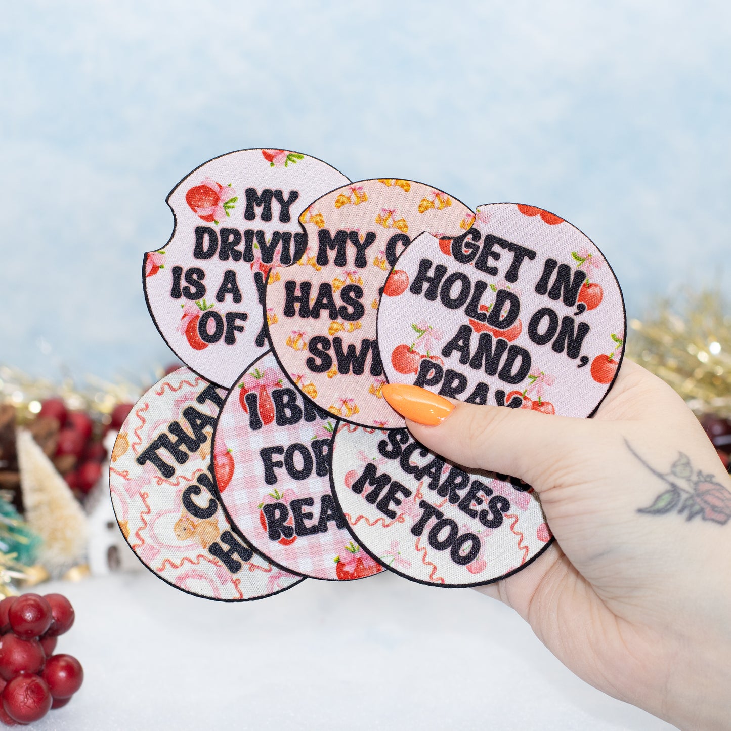 A hand holds novelty coasters with black text and red fruit illustrations on a pink gingham background. The background is a softly blurred festive holiday scene.