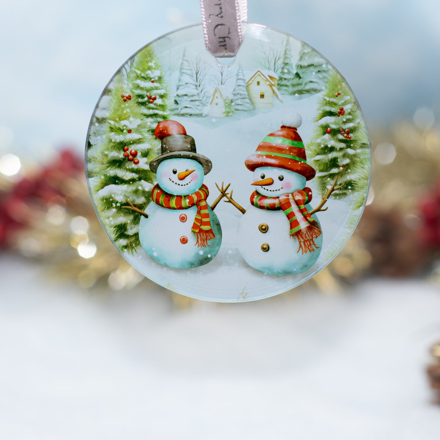 A round glass Christmas ornament featuring two snowmen in a snowy winter village scene, hanging by a ribbon against a softly blurred festive background.