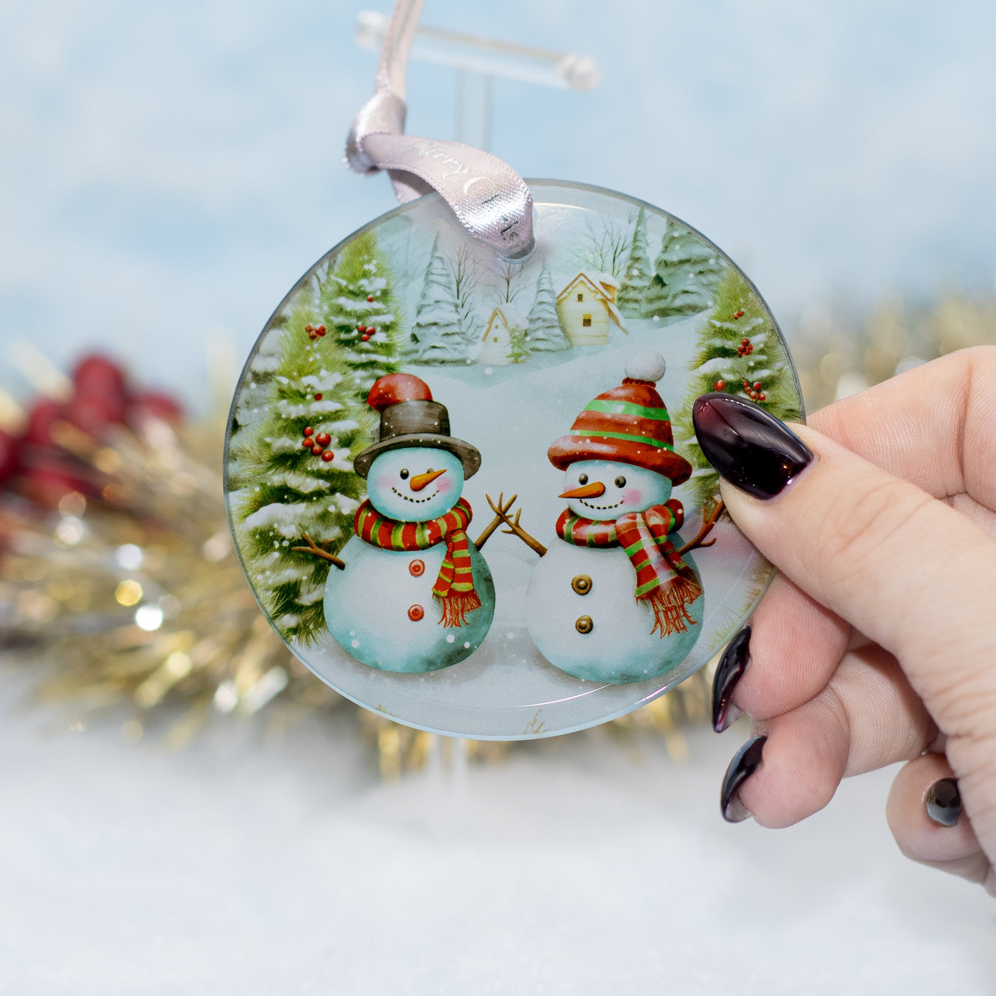 A hand holds a round glass Christmas ornament featuring two snowmen in winter clothing, with a blurred festive background.