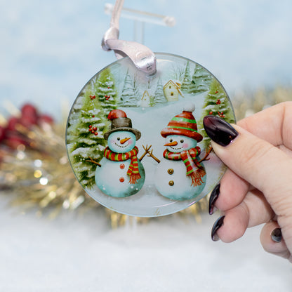 A hand holds a round glass Christmas ornament featuring two snowmen in winter clothing, with a blurred festive background.