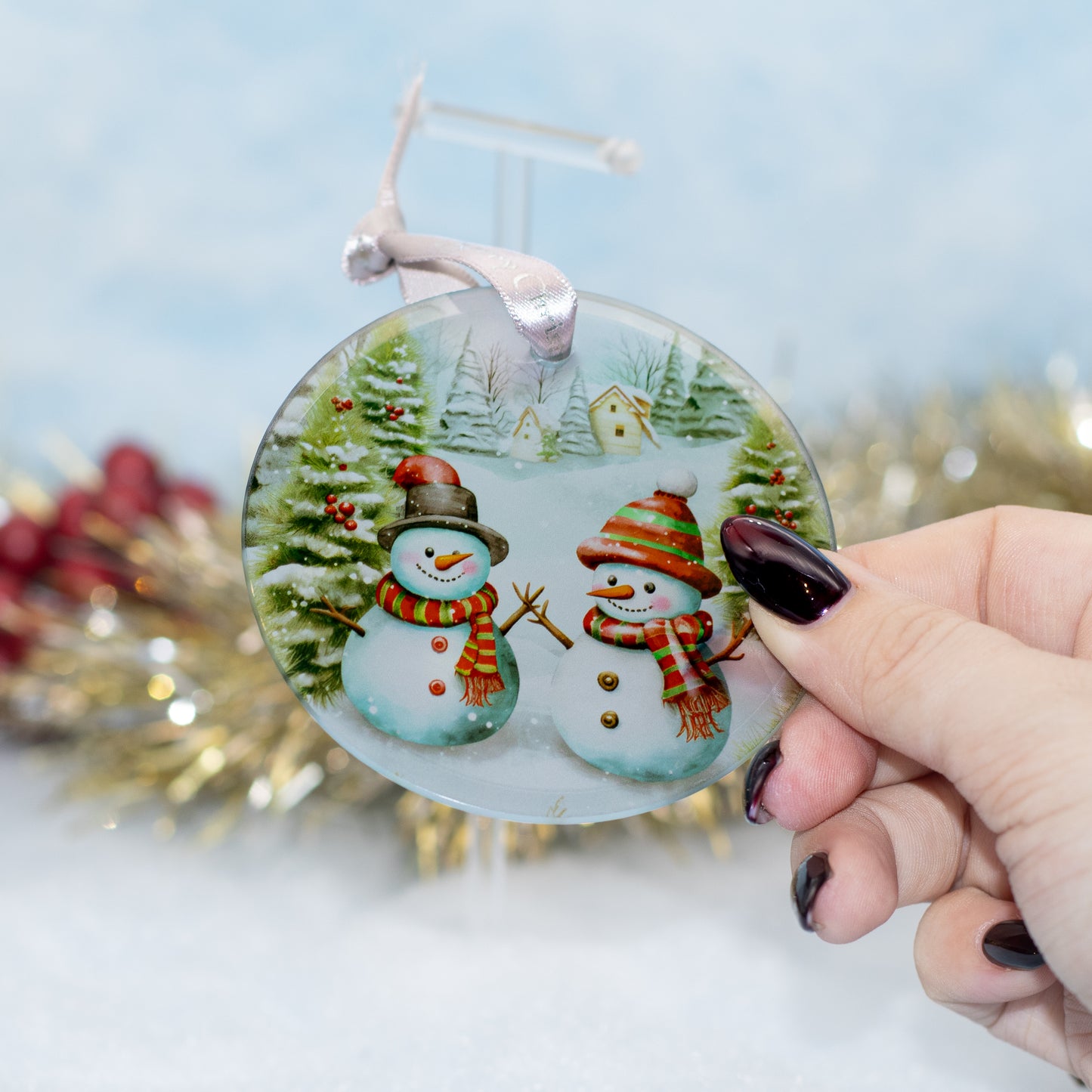 A hand holds a round glass Christmas ornament featuring two snowmen in a snowy winter village. The ornament has a silver hanging loop and is shown against a softly blurred festive background with gold tinsel and red berries.