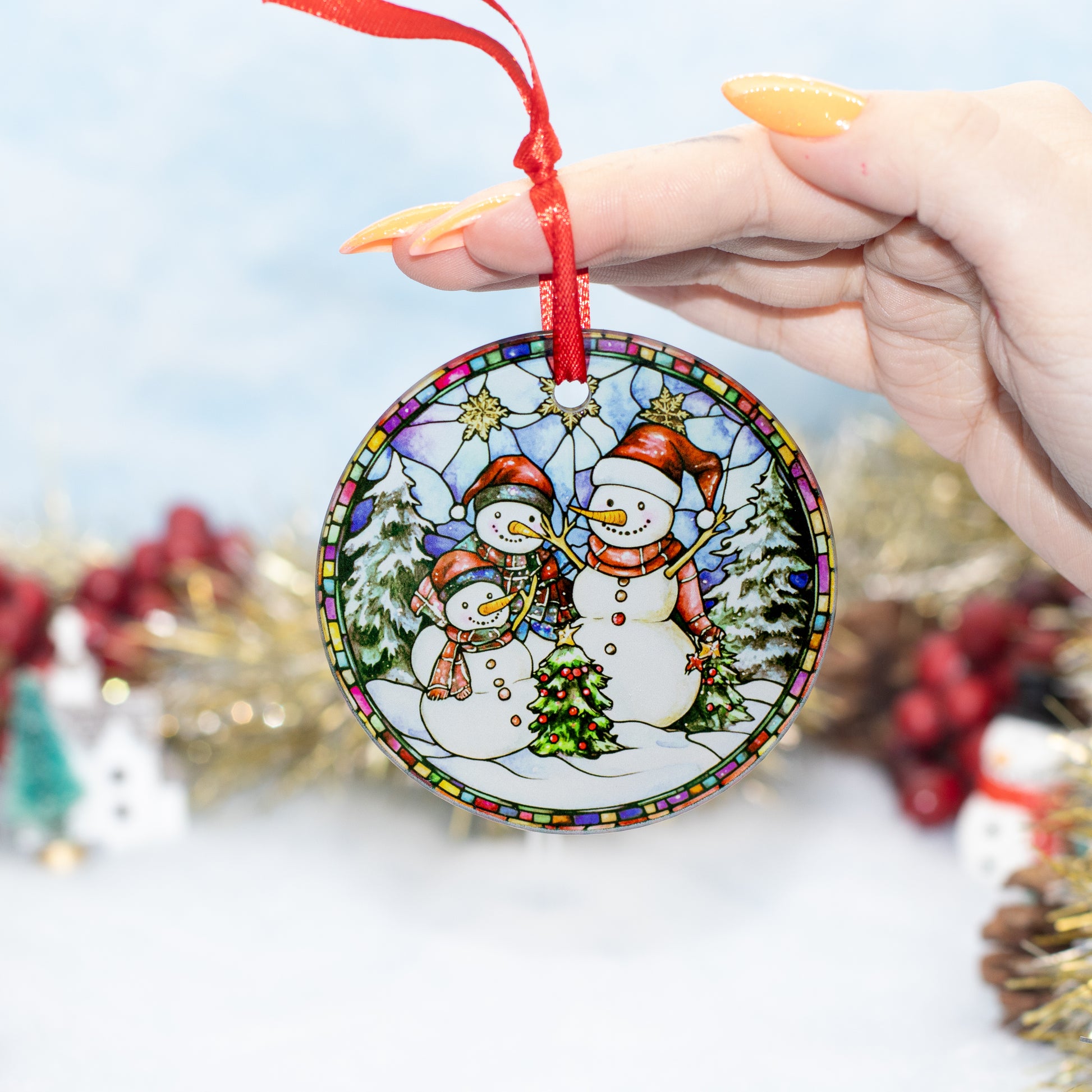 A hand holds a round ceramic Christmas ornament featuring three snowmen in a winter scene with a red hanging ribbon. The background is a soft, out-of-focus holiday setting with gold tinsel and red berries.