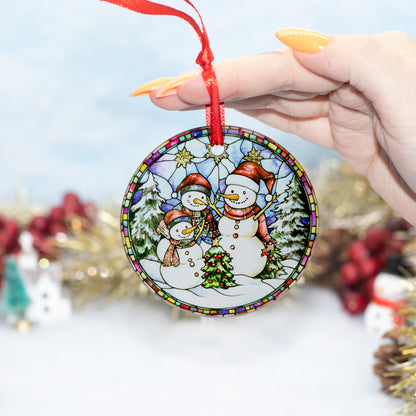 A hand holds a round ceramic Christmas ornament featuring three snowmen in a winter scene with a red hanging ribbon. The background is a soft, out-of-focus holiday setting with gold tinsel and red berries.