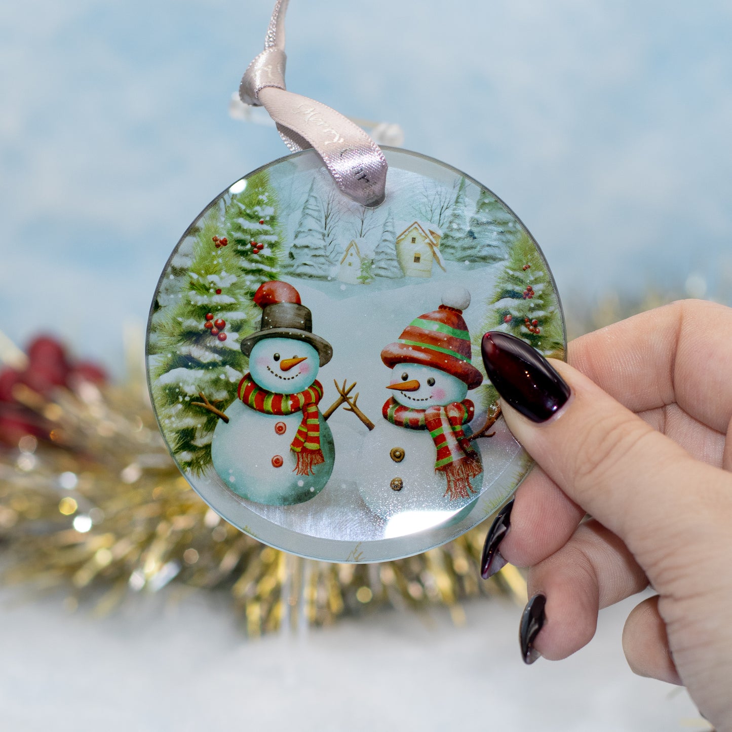 A round glass Christmas ornament featuring two snowmen in winter attire, held by a hand with dark manicured nails against a soft, blurred festive background.