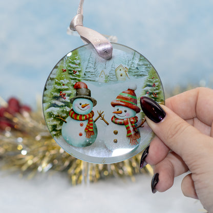 A round glass Christmas ornament featuring two snowmen in winter attire, held by a hand with dark manicured nails against a soft, blurred festive background.