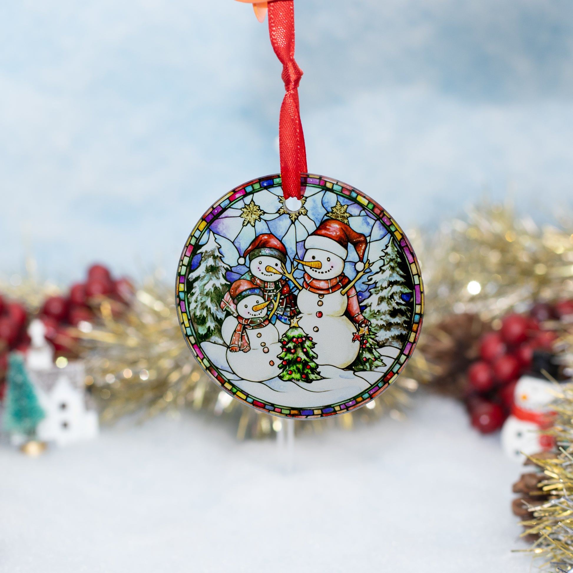 A round Christmas ornament featuring three snowmen in a winter scene, hanging from a red ribbon against a softly blurred festive background with tinsel and berries.