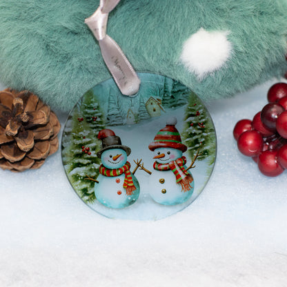 A round Christmas ornament featuring two snowmen in winter clothing, displayed on a snowy surface with a green fuzzy blanket, pinecone, and red berries in the background.