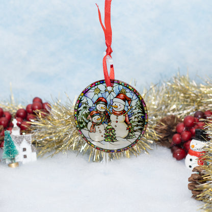 A round Christmas ornament featuring a stained-glass style illustration of two snowmen, hanging by a red ribbon in a snowy decorative setting with tinsel and berries.